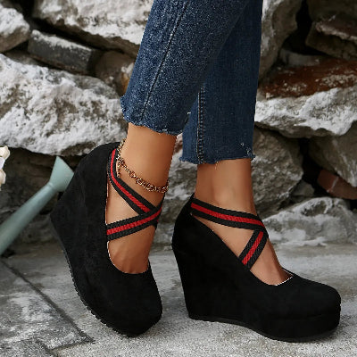 Black wedge sandals with red and black straps worn by a person against a stone wall background.