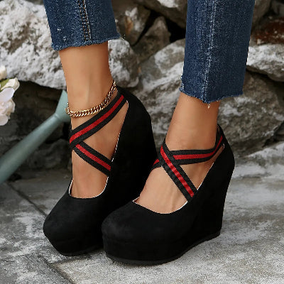 Black wedge sandals with red straps worn by a person against a stone wall background