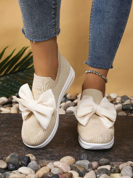 Beige slip-on shoes with large bows worn with rolled-up jeans on a pebbly surface.